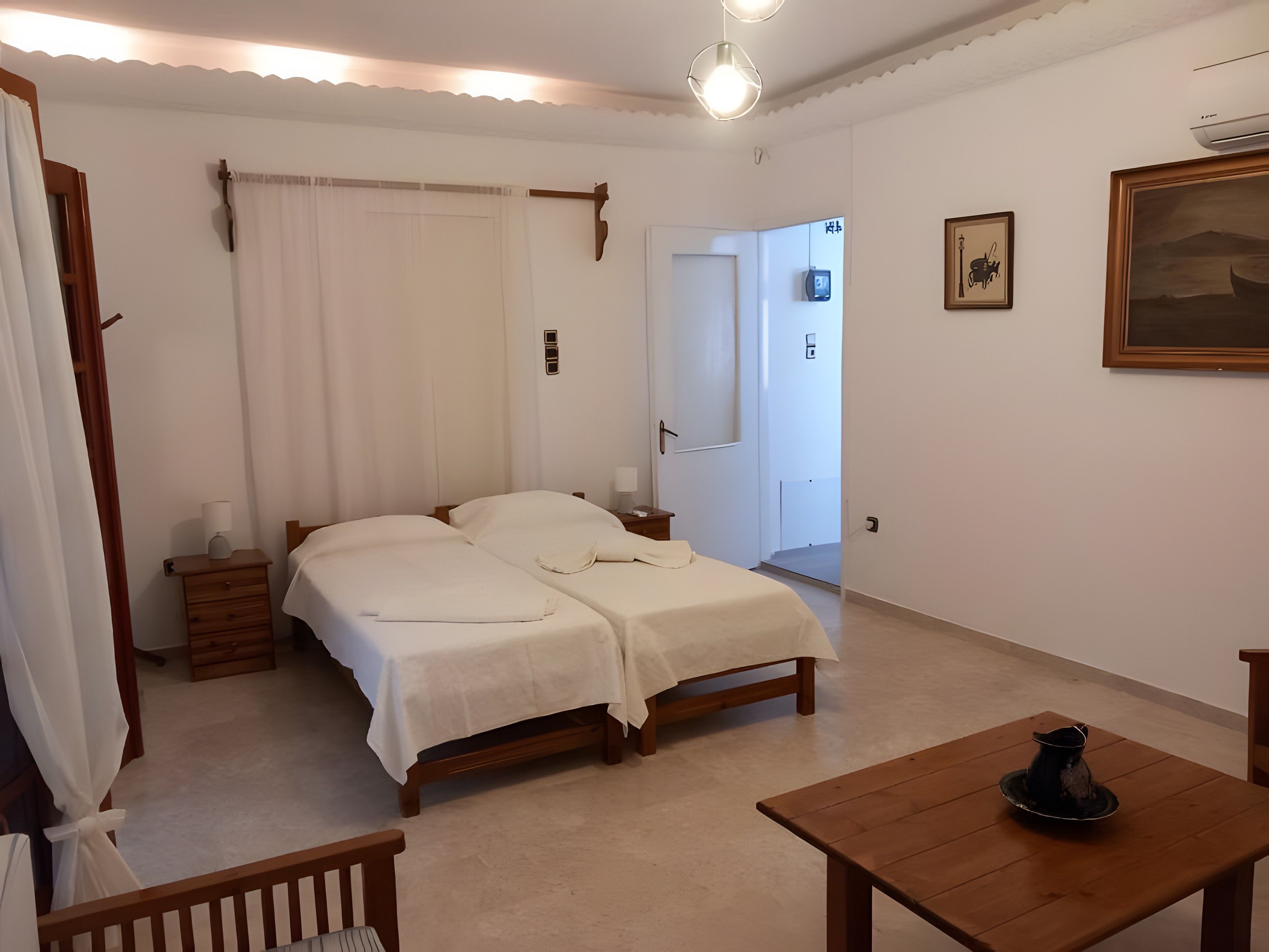 Une chambre avec deux lits, une table de chevet en bois, une commode, une chaise en bois, une table avec une carafe, et une porte donnant sur une autre pièce.