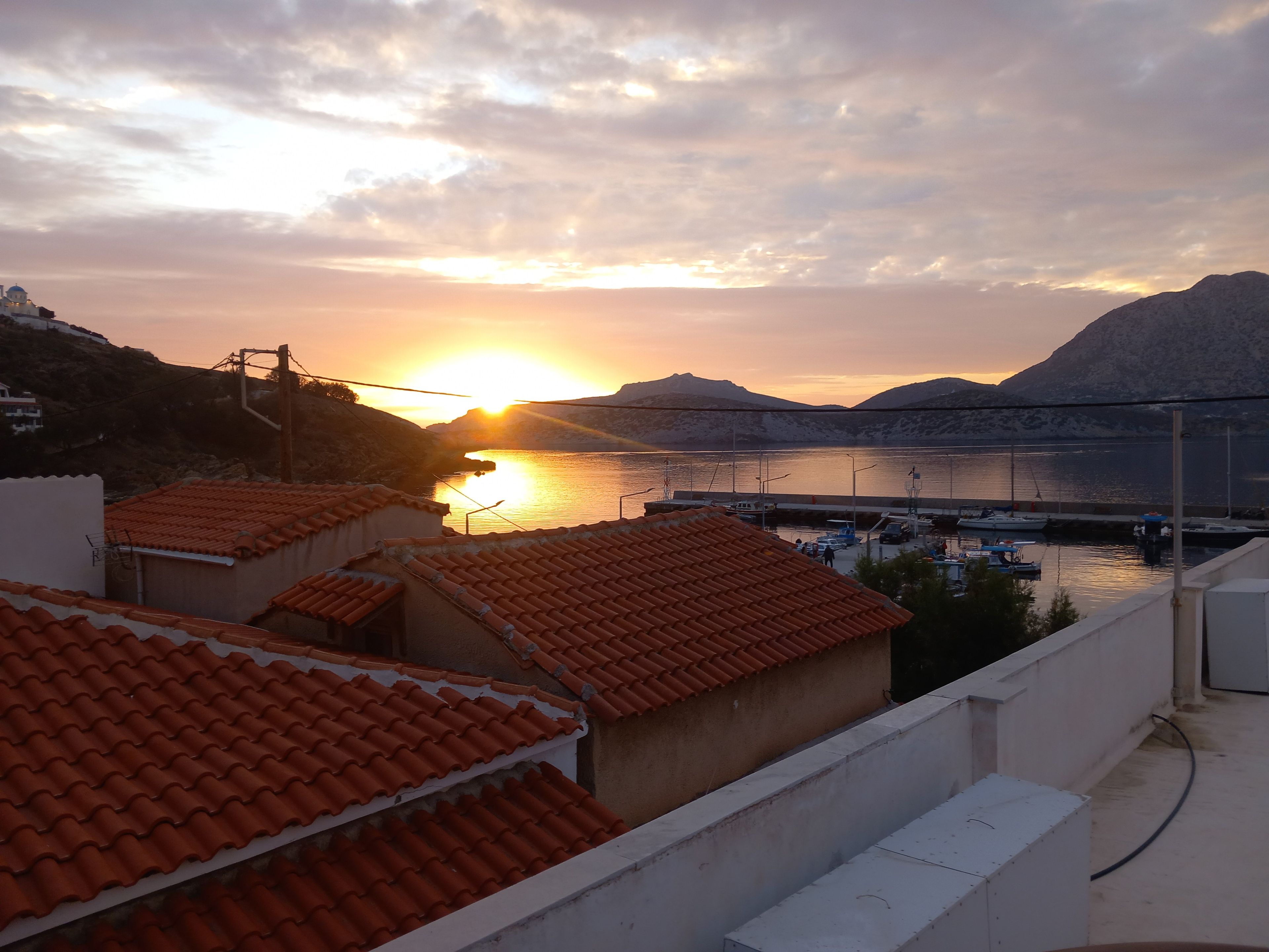 A coastal view with a sunset, buildings with red-tiled roofs, and boats docked at a marina.