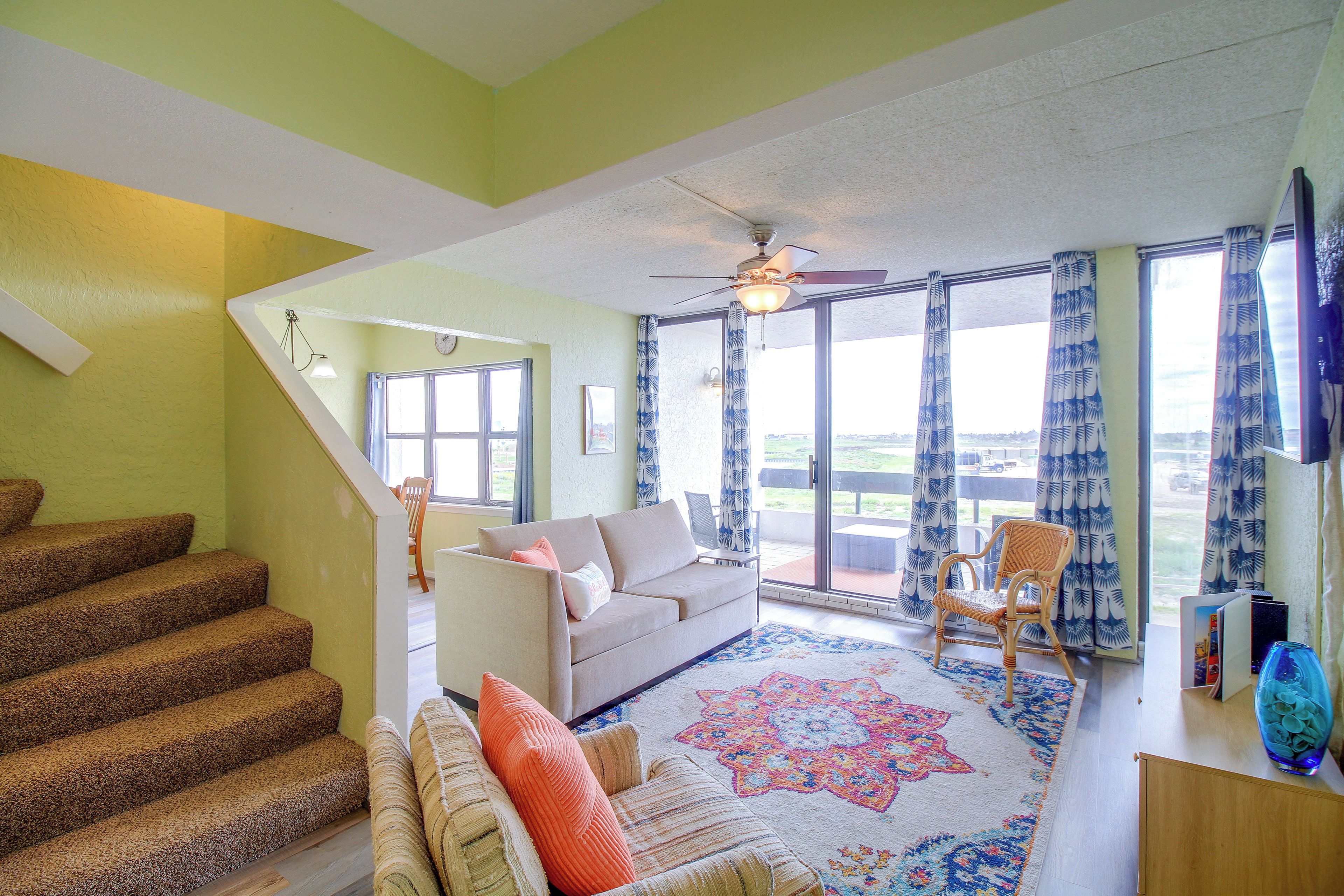 A living room with a staircase, a sofa, a patterned rug, and a view through large windows.