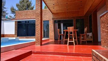 A modern outdoor patio with a pool, wooden furniture, and a brick exterior.