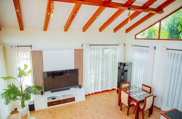 A living room with a flat-screen TV, wooden beams, a dining table with chairs, and a potted plant.