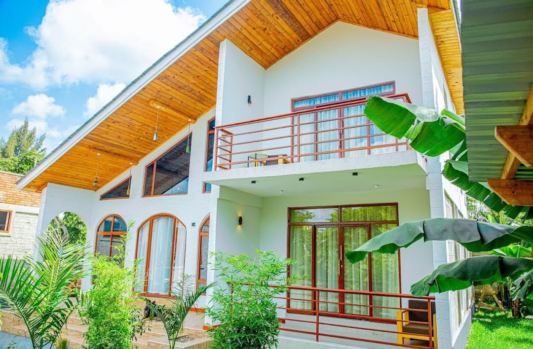 A two-story building with a balcony, large windows, and a garden with tropical plants.