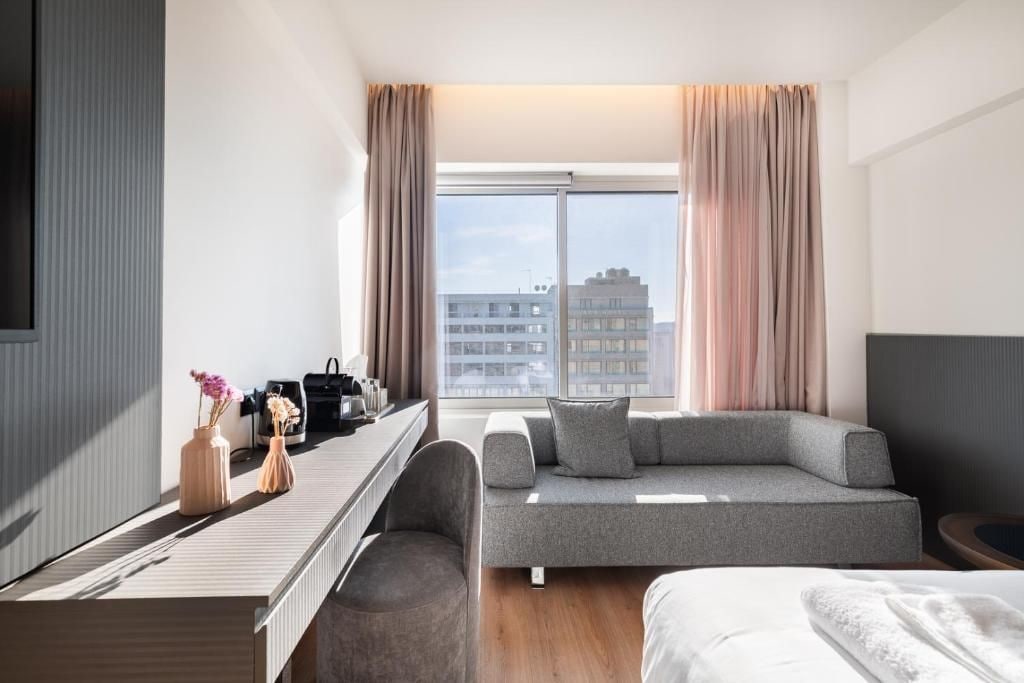 A modern hotel room with a sofa, a bed, a desk with a computer, and a large window with curtains.