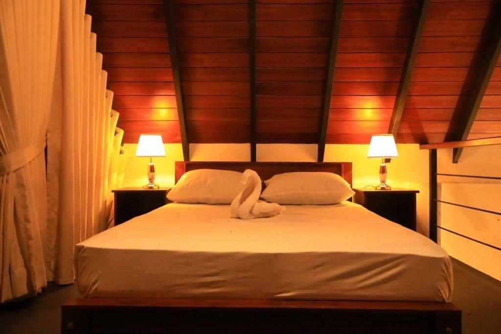 Ein Schlafzimmer mit Holzbalkendecke, zwei Nachttischlampen, einem Bett mit weißer Bettwäsche und einer Schwan-Form-Kissenanordnung.