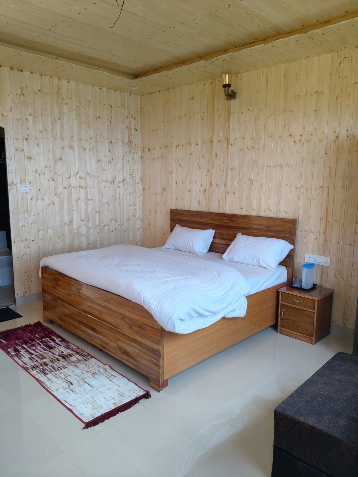 A wooden-paneled bedroom with a wooden bed, a nightstand, and a red and white rug.