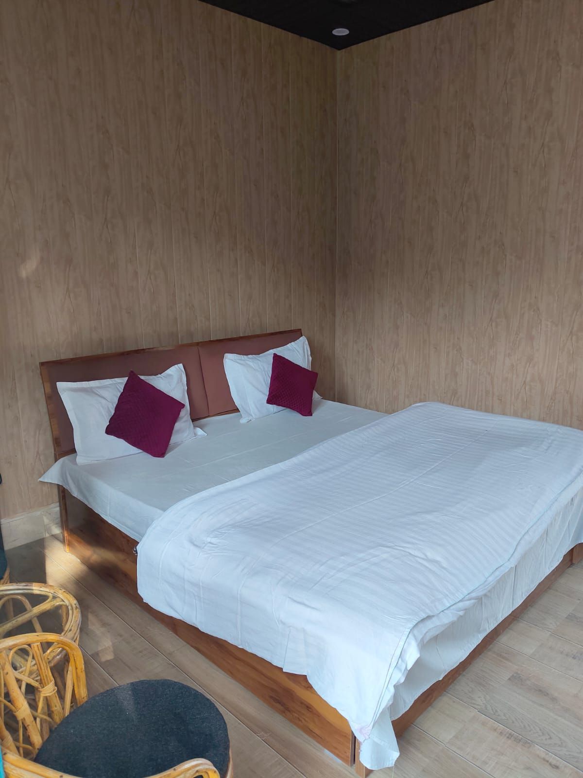 Una cama individual con cabecera de madera, ropa de cama blanca y dos almohadas moradas. En primer plano se ve una mesa redonda con tapa negra.
