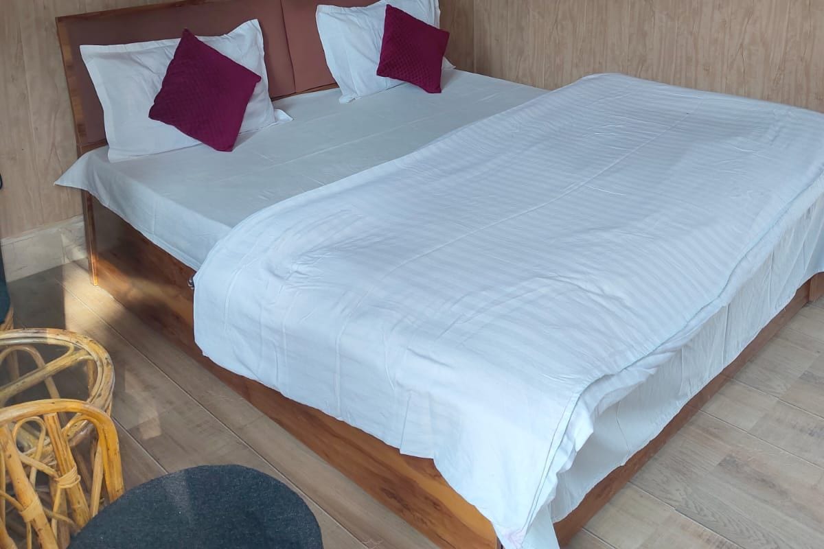Yhden hengen sänky, jossa on puinen pääty, valkoiset vuodevaatteet ja kaksi violettia tyynyä. Kuvan etualalla näkyy pyöreä pöytä, jonka kansi on musta.