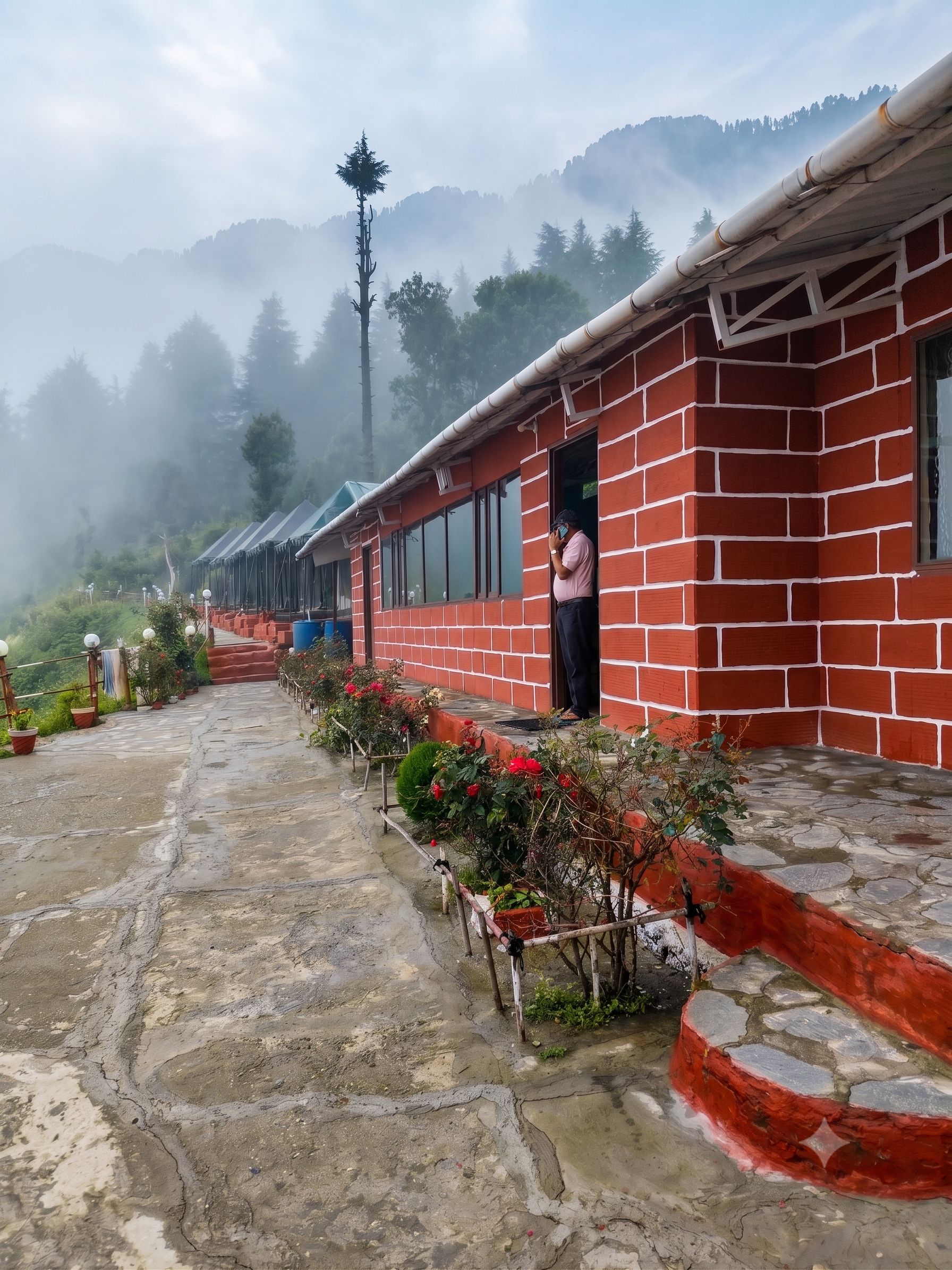 A row of red brick buildings with a person standing at the entrance, a stone-paved pathway, and a mountainous backdrop.