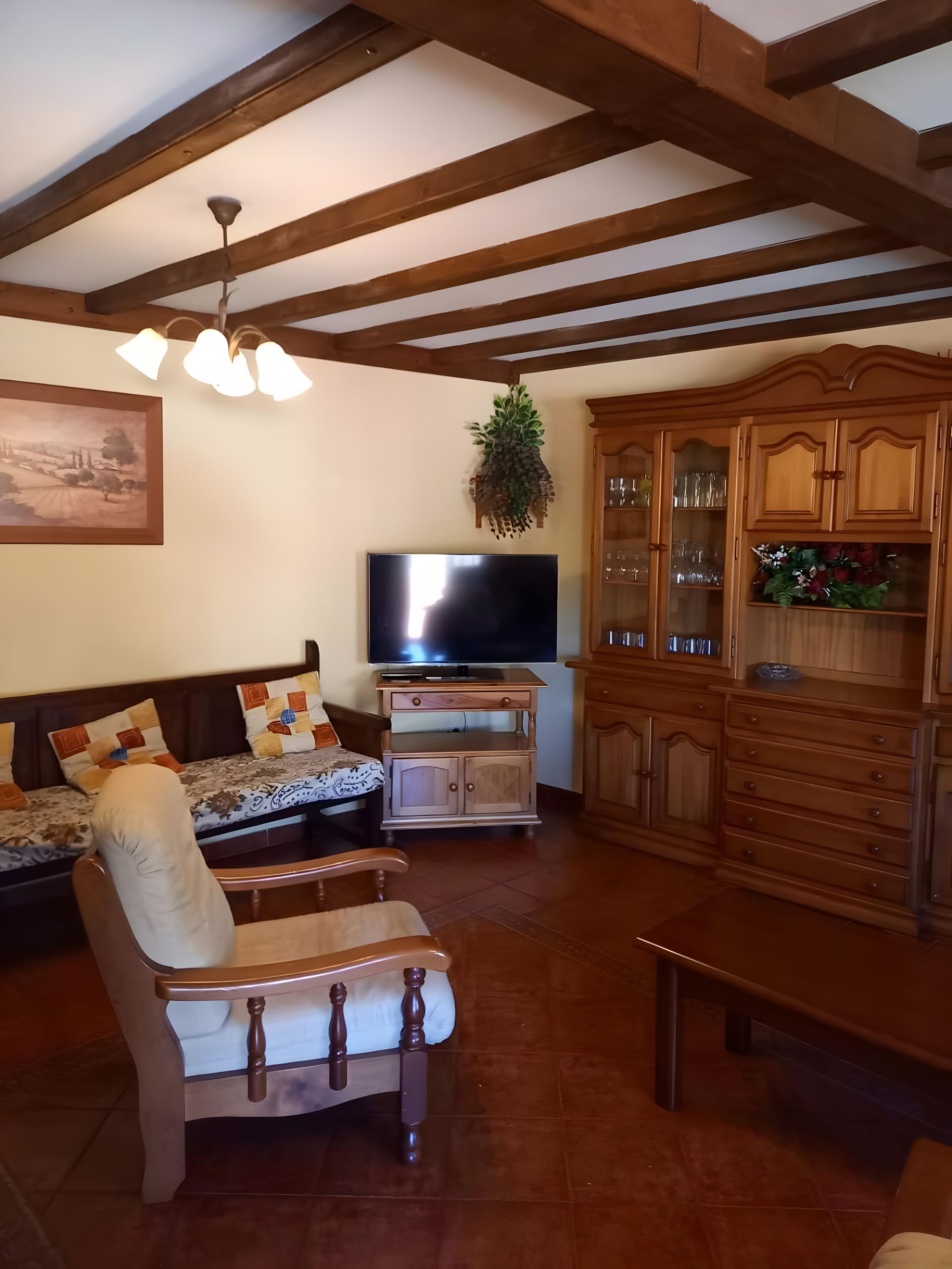 Een woonkamer met een bank, een stoel, een televisie, houten kasten en een plafond met zichtbare balken.