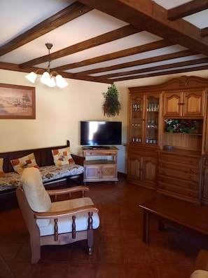 A living room with a sofa, a chair, a television, wooden cabinets, and a ceiling with exposed beams. - Country House 'Casa Rural La Cerra' with Mountain View, Shared Garden and Wi-Fi (Navalguijo)