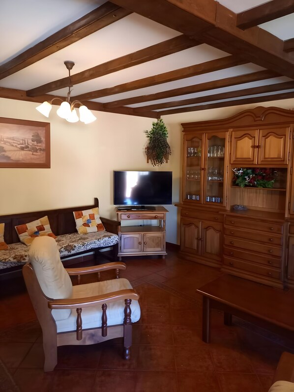 Un salon comprenant un canapé, un fauteuil, un téléviseur, des meubles en bois et un plafond avec des poutres apparentes.