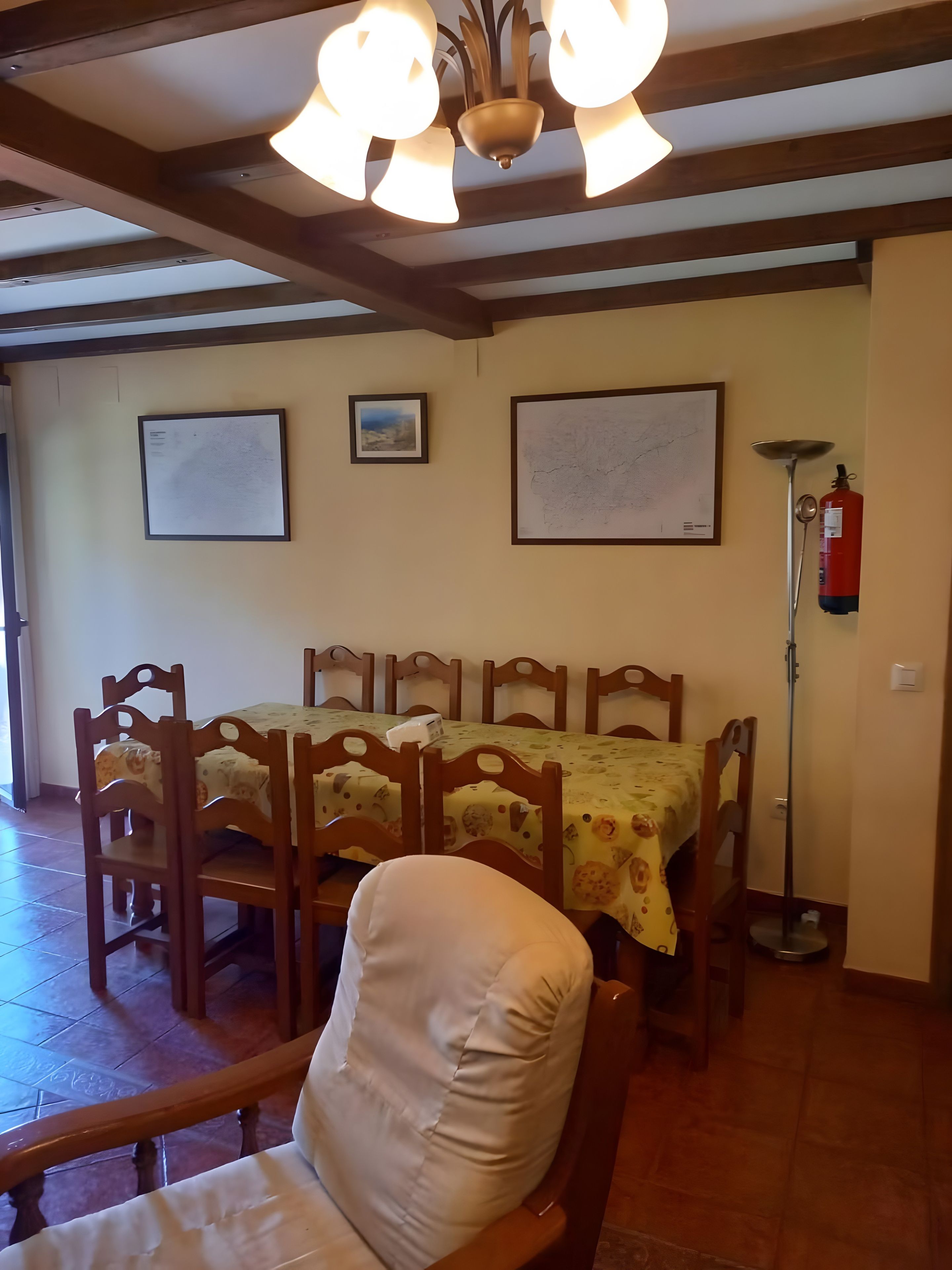 Een eethoek met houten stoelen en een tafel met een tafelkleed met bloemenprint. Er hangen twee ingelijste foto's aan de muur, een brandblusser en een vloerlamp.