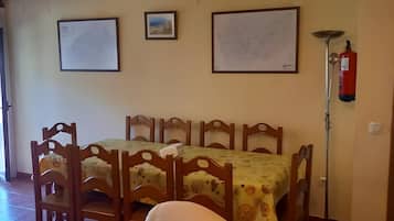 Een eethoek met houten stoelen en een tafel met een tafelkleed met bloemenprint. Er hangen twee ingelijste foto's aan de muur, een brandblusser en een vloerlamp.