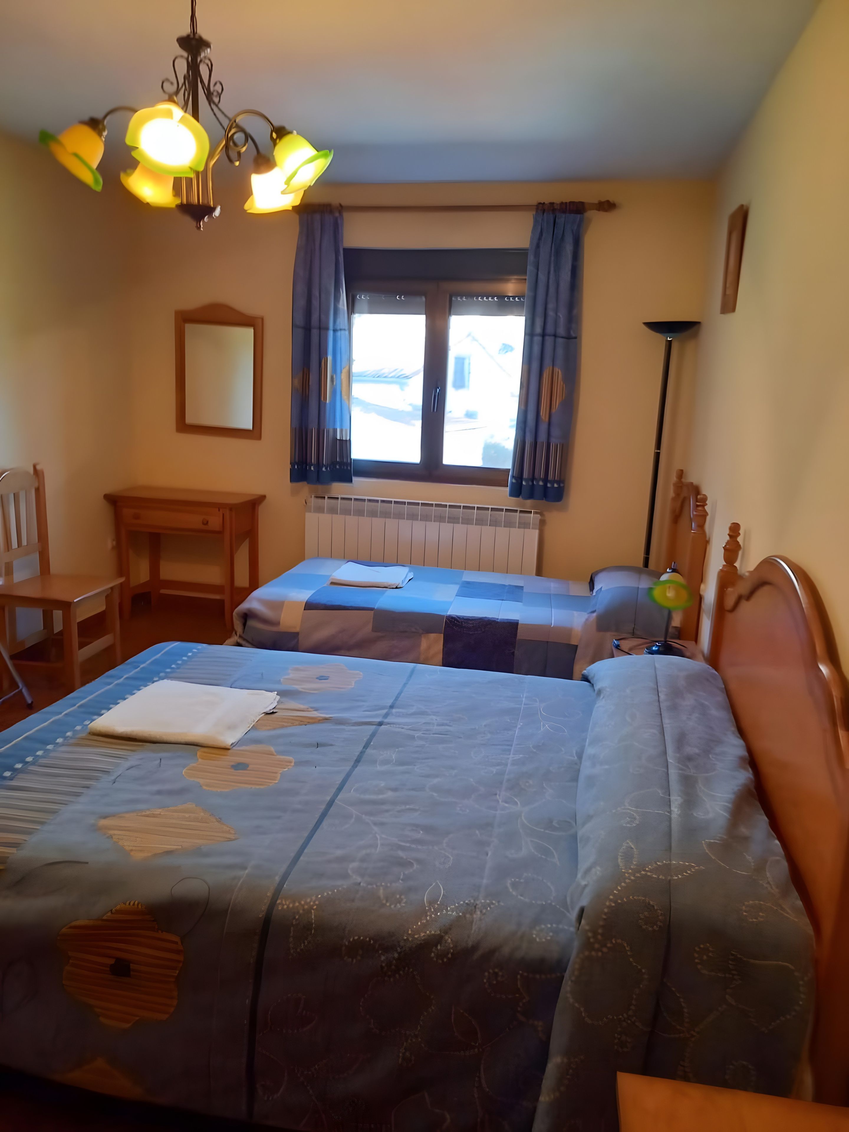 Une chambre avec deux lits, un bureau, une chaise, une fenêtre avec des rideaux et un luminaire suspendu.