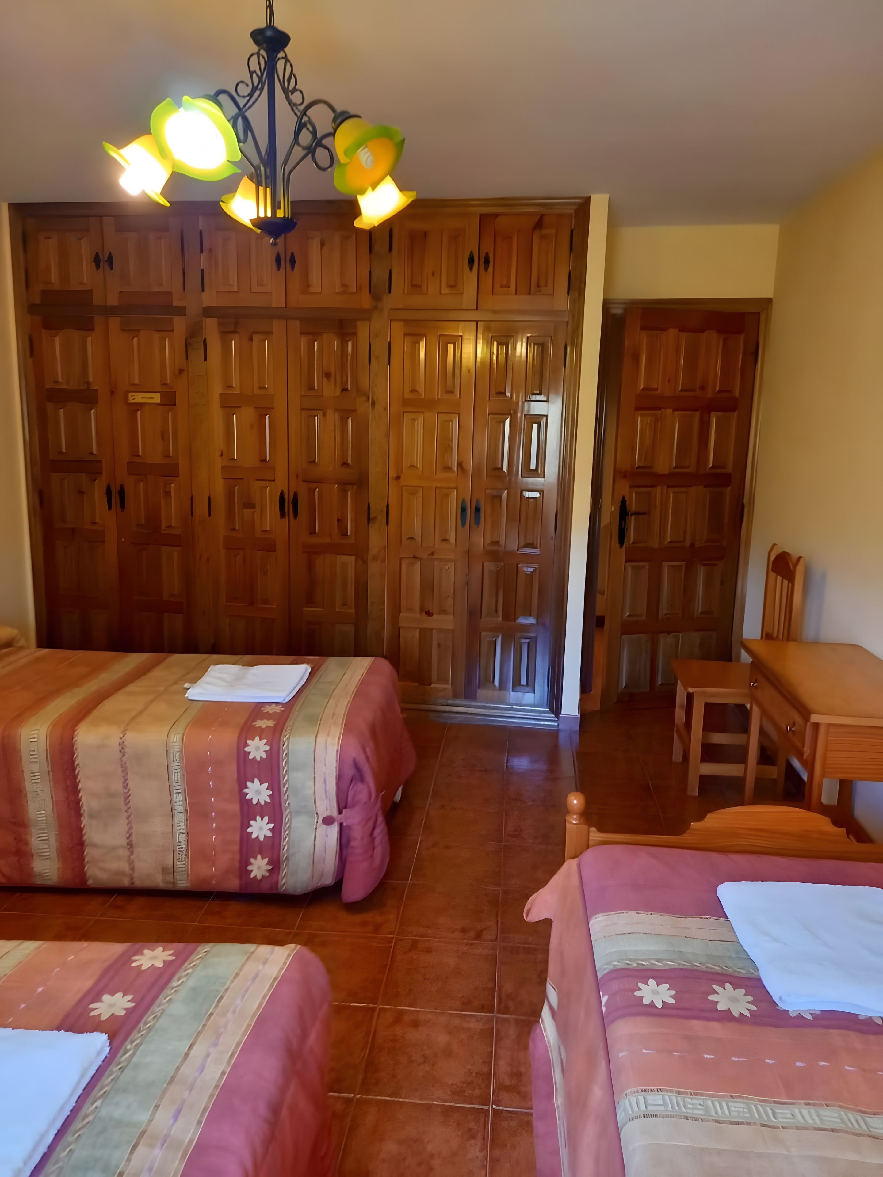 A hotel room with two beds, a wooden wardrobe, a dining table, and a chandelier.
