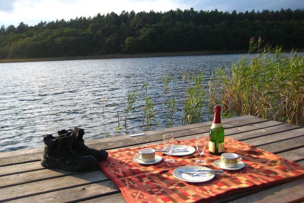 Un montaje para picnic en una terraza de madera junto a un lago, con una botella de champán, platos, vasos y un par de botas.
