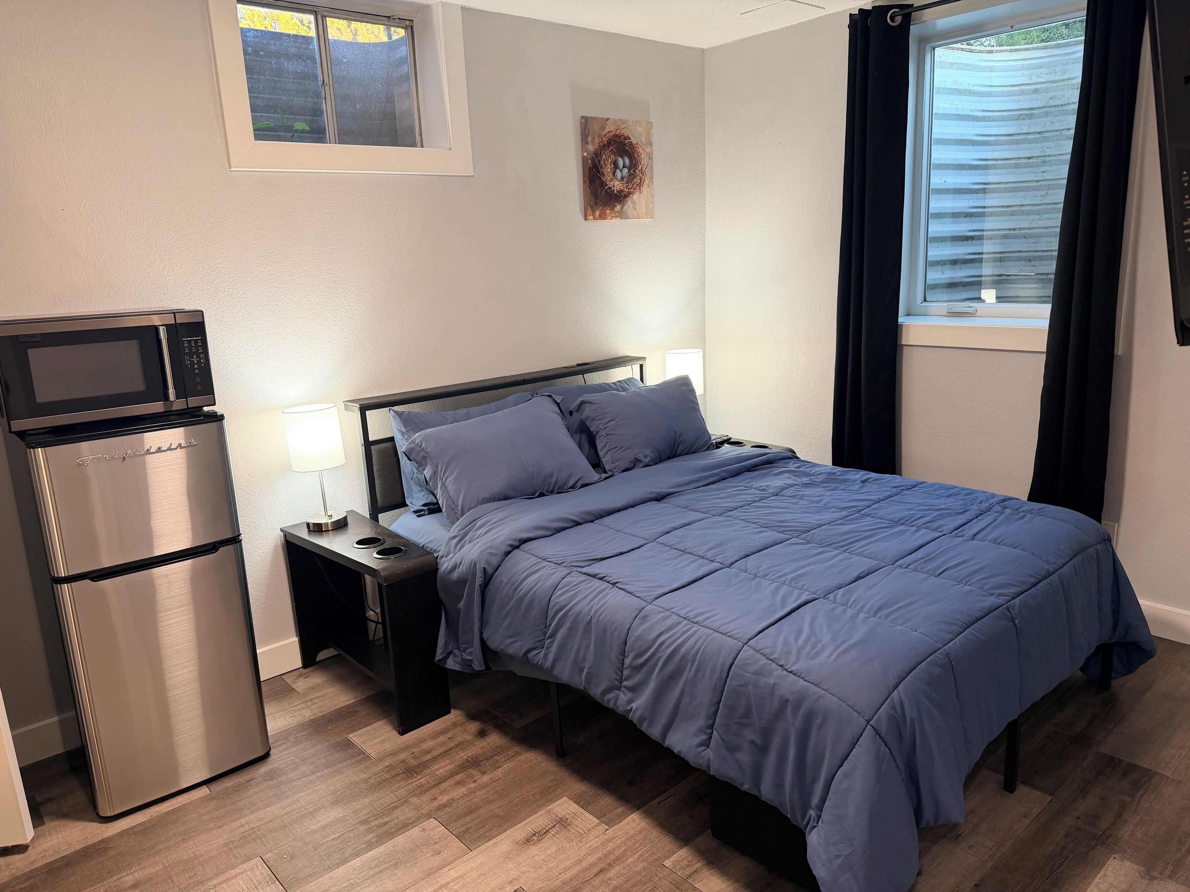 A bedroom with a bed, a nightstand, a lamp, a built-in microwave, and a refrigerator.