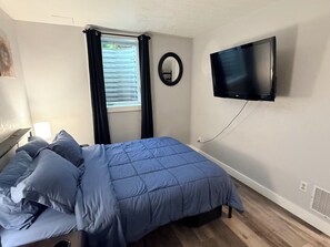 Ein Schlafzimmer mit einem Bett, einem an der Wand montierten Fernseher, einem Fenster mit Vorhängen und einem Nachttisch mit einer Lampe.