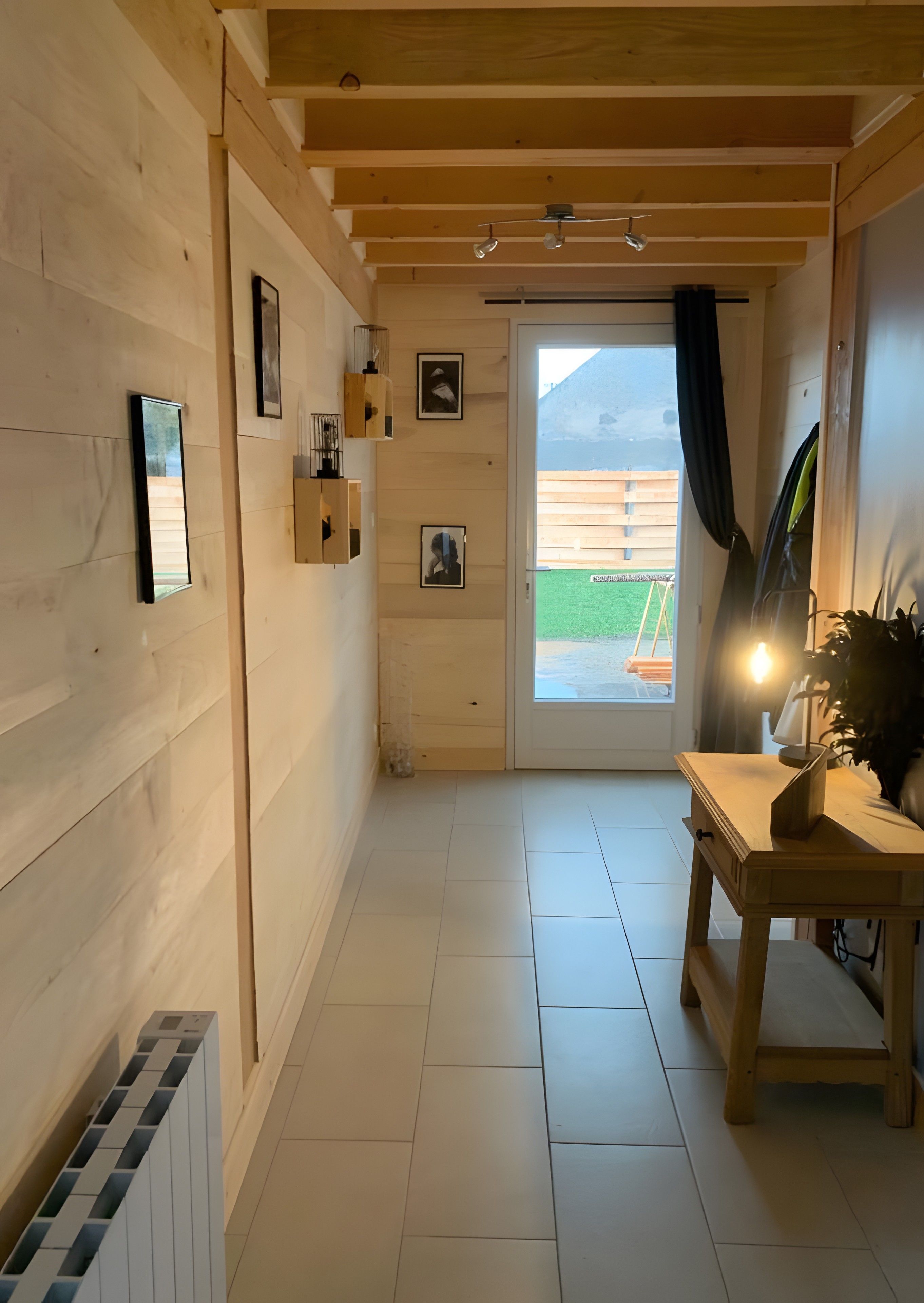 Un couloir aux murs en bois, un sol carrelé, une table avec une lampe et une porte donnant sur l’extérieur.