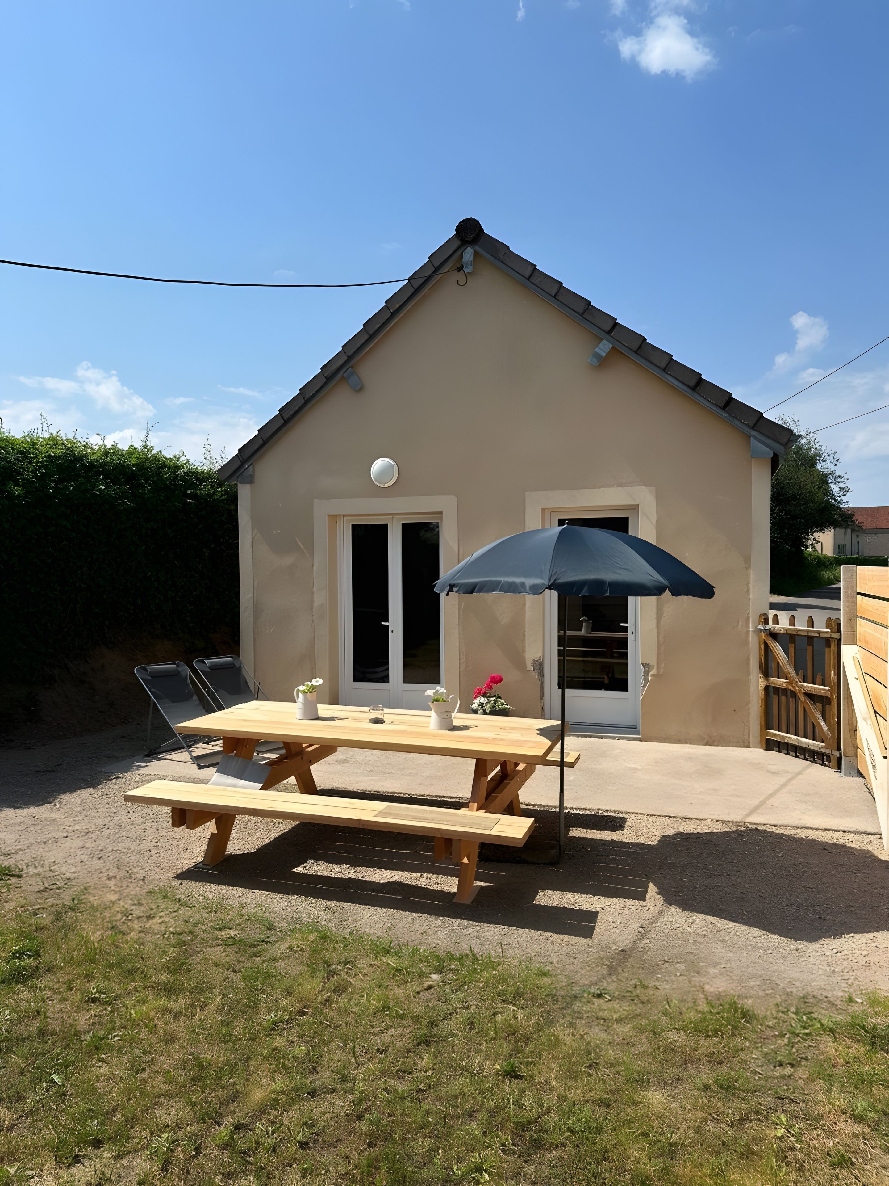 A house with a patio area featuring a wooden table and chairs, a black umbrella, and a small garden.