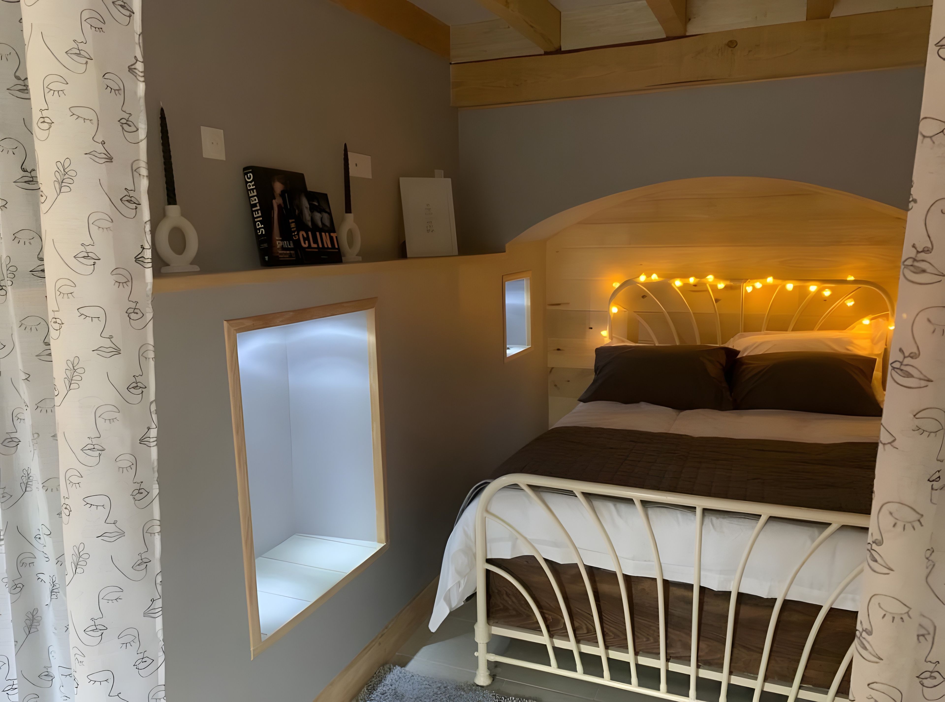A bedroom with a white metal bed frame, a built-in shelf with books and a lamp, a small window, and string lights on the headboard.