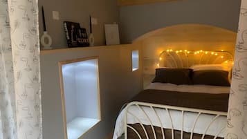 A bedroom with a white metal bed frame, a built-in shelf with books and a lamp, a small window, and string lights on the headboard.