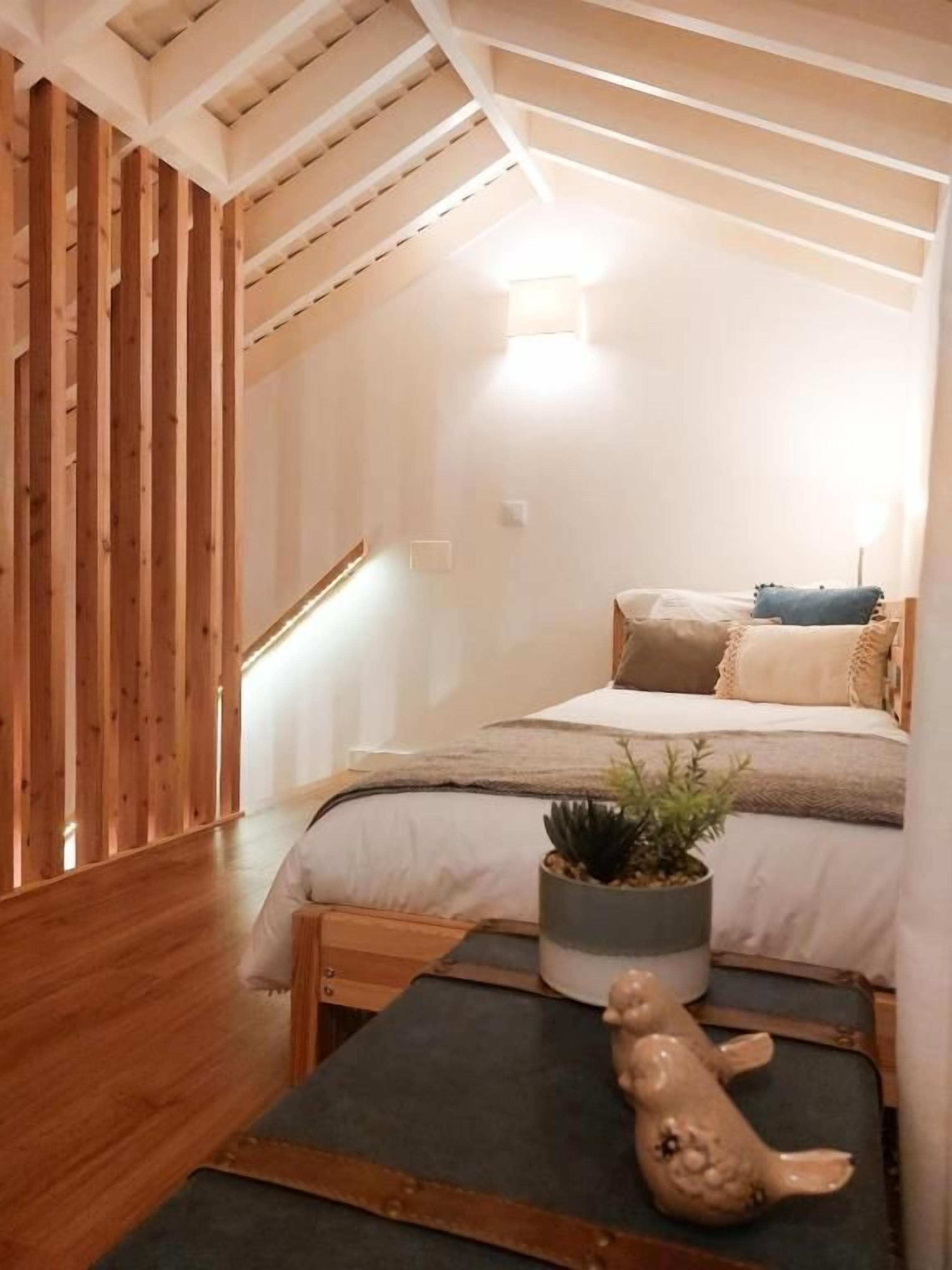 Un dormitorio con techo inclinado, una cama con almohadas, una planta pequeña y un objeto decorativo sobre un banco.