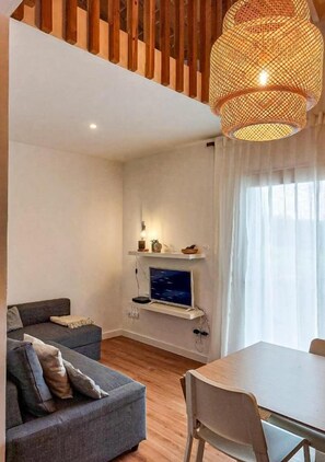 Ein modernes Wohnzimmer mit grauem Sofa, einem Esstisch, einem Fernseher an einer Wandhalterung und einer großen gewebten Leuchte.