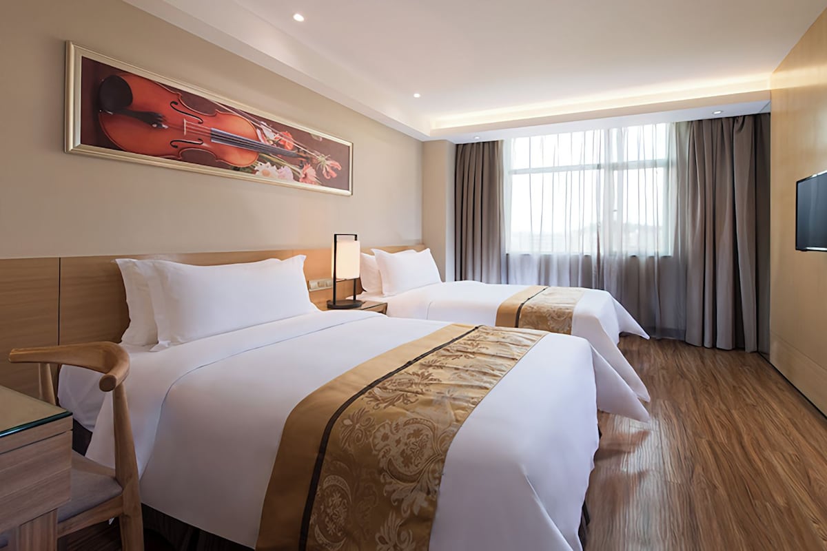 Een hotelkamer met twee bedden, een bureau, een stoel, een schilderij aan de muur en een raam met gordijnen.
