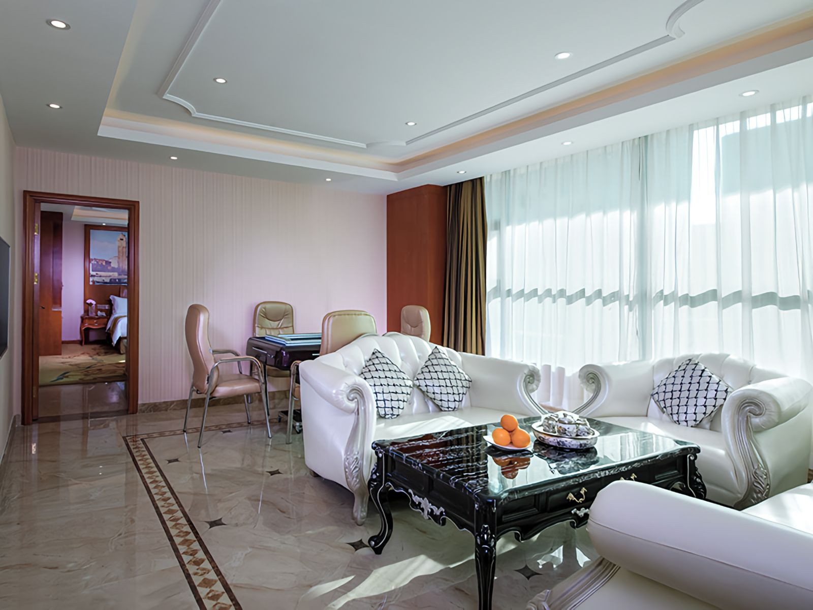 A modern hotel room with a white sofa set, a glass coffee table, and a dining area with chairs.