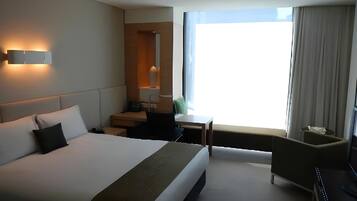 Ein Hotelzimmer mit einem großen Bett, einem Schreibtisch, einem Stuhl, einem kleinen Tisch, einer Lampe und einem Fenster mit Vorhängen.
