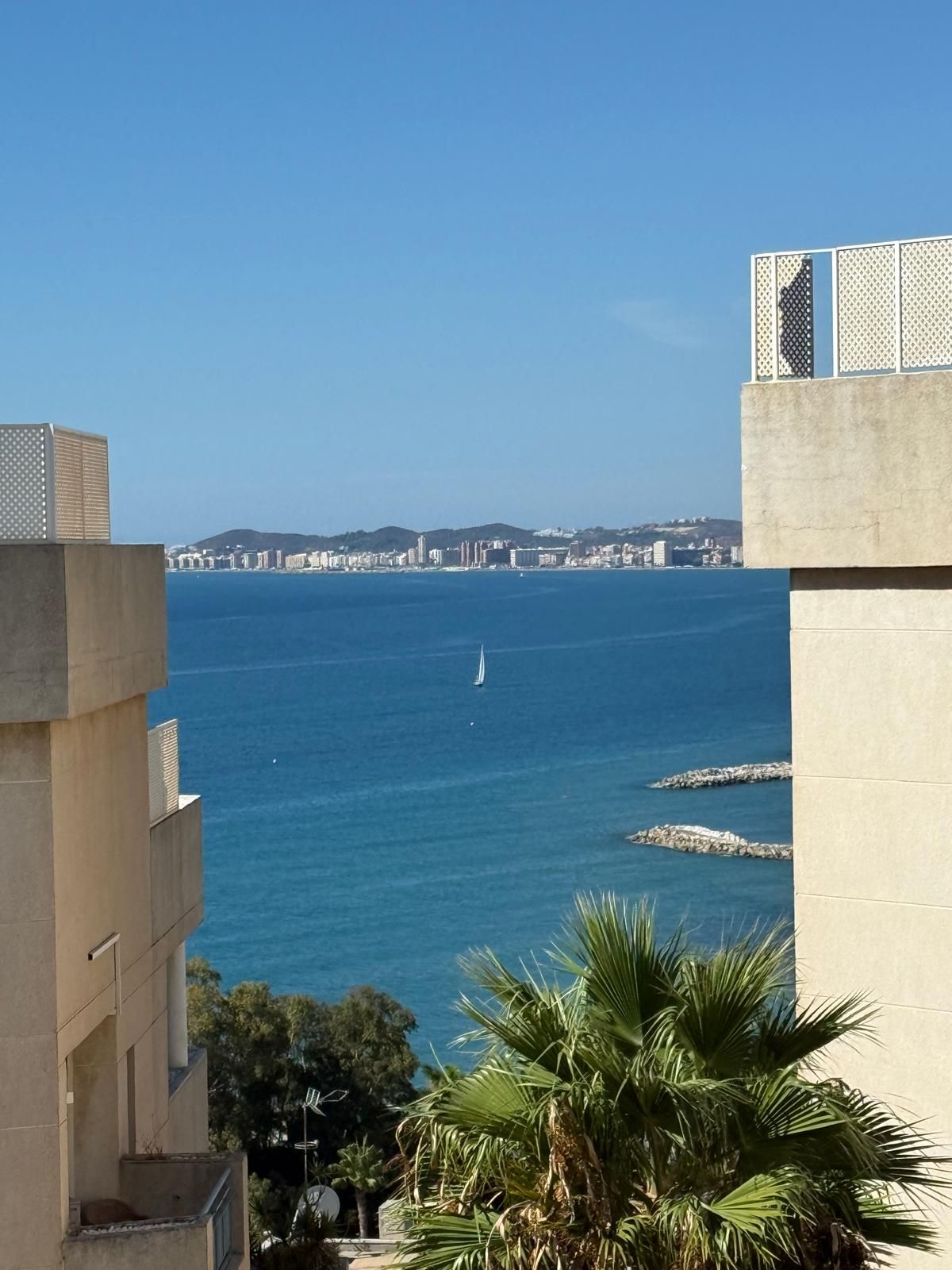 Un paisaje urbano costero con vista despejada al mar, un velero y edificios en la distancia.