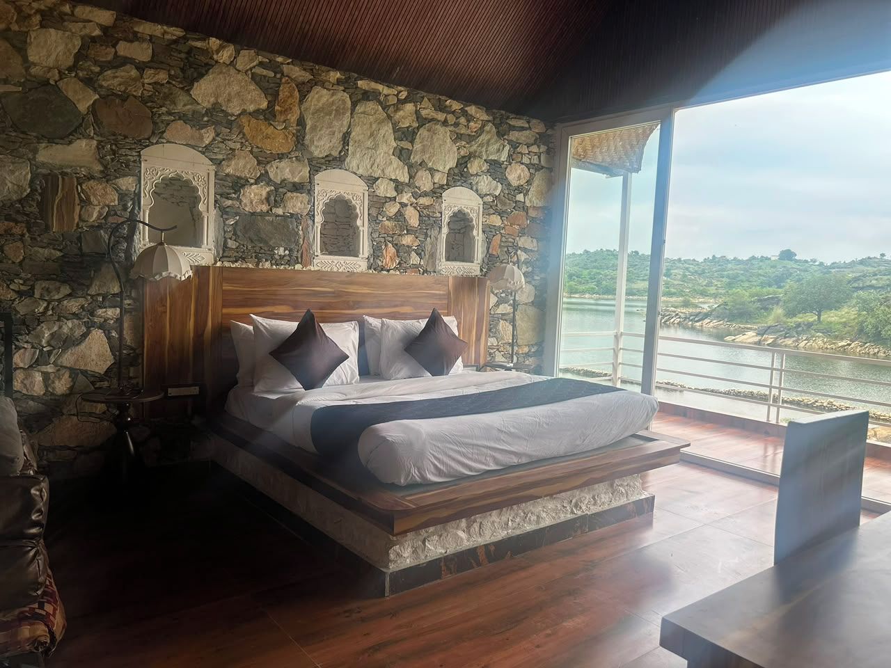 A bedroom with a stone wall, a wooden bed, and a view of a body of water.