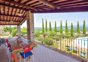 Une terrasse extérieure couverte, avec une table et des chaises, donnant sur la campagne et à proximité d’une piscine.