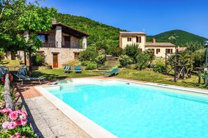 Un espace piscine avec une piscine à l’eau d’un bleu limpide, entourée d’un bâtiment en pierre doté d’un balcon, et un bâtiment plus petit en arrière-plan.