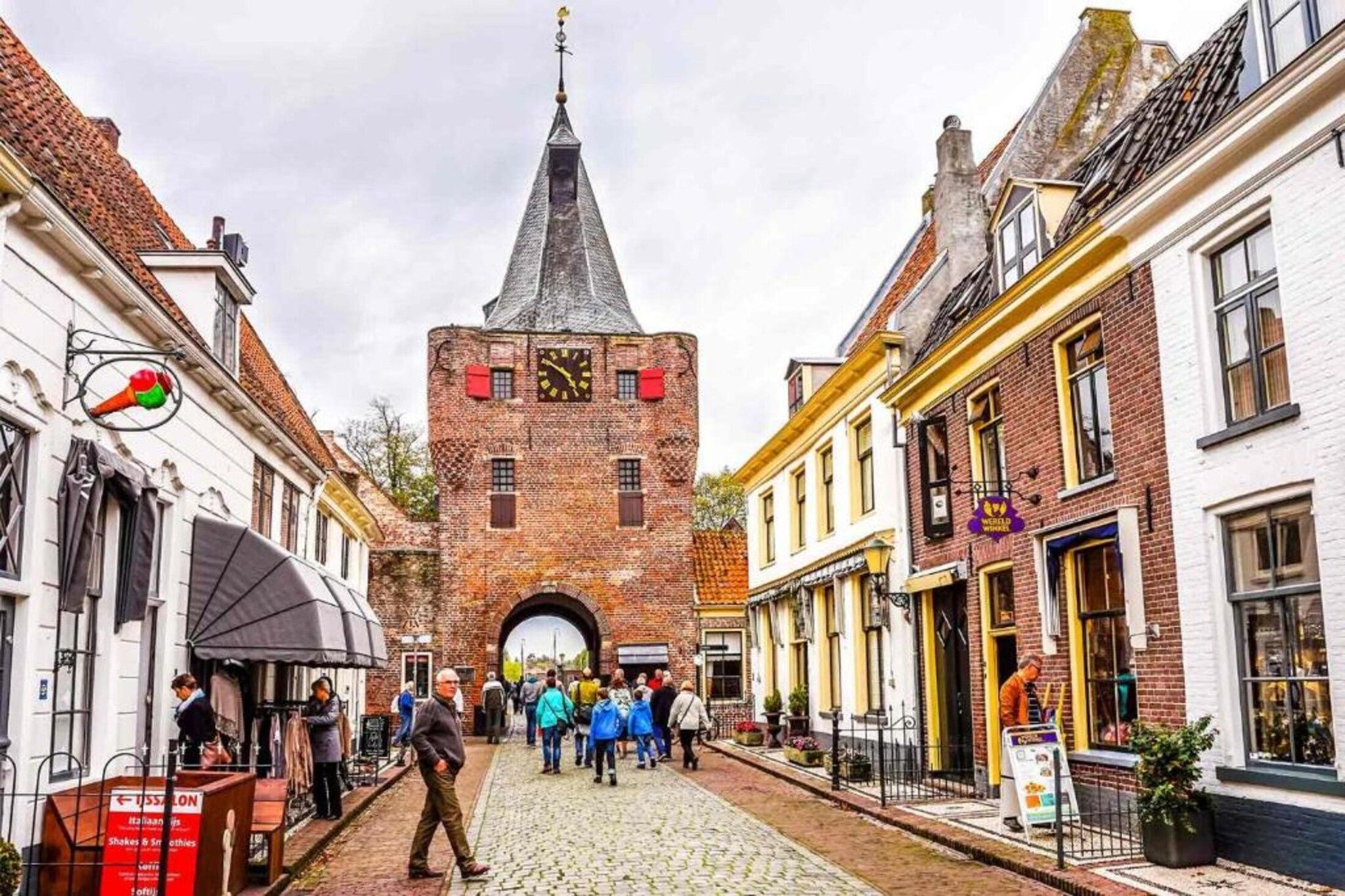 Eine gepflasterte Straße mit einem markanten Backsteinturm mit einer Uhr, flankiert von traditionellen Gebäuden mit Geschäften und Außenplätzen.
