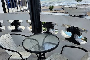 Ein Balkon mit einem runden Tisch und zwei weißen Stühlen, der Blick auf einen Strand mit einem Boot und Personen in der Ferne gewährt.