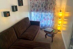 Ein Wohnzimmer mit einem braunen Sofa, einem kleinen runden Tisch mit einem Buch und einer Fernbedienung sowie einer Wandlampe.