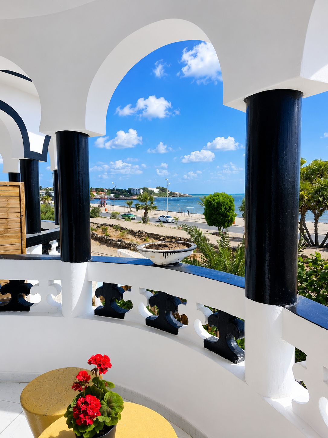 Un balcon donnant sur une plage et la mer, agrémenté d’une balustrade blanche, d’un pot de fleurs et d’un banc en bois.