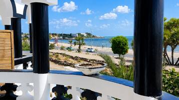 Un balcon donnant sur une plage et la mer, agrémenté d’une balustrade blanche, d’un pot de fleurs et d’un banc en bois.