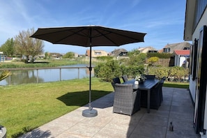 Une terrasse extérieure avec une table et des chaises, un grand parasol, et une vue sur un lac et des maisons en arrière-plan.