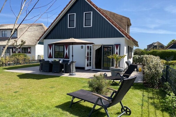 Une maison à deux étages, à l’extérieur sombre, avec un toit de chaume et un patio aménagé avec du mobilier d’extérieur.