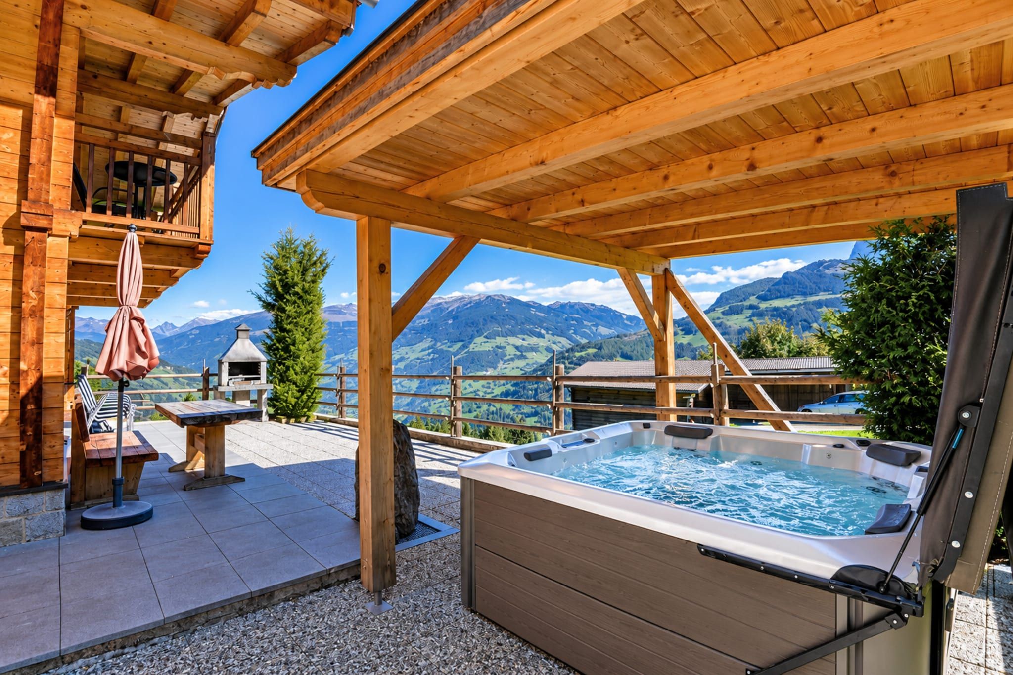 Un espace extérieur aménagé avec un bain à remous en bois, offrant une vue sur les montagnes, ainsi qu’un barbecue et un coin salon abrité.