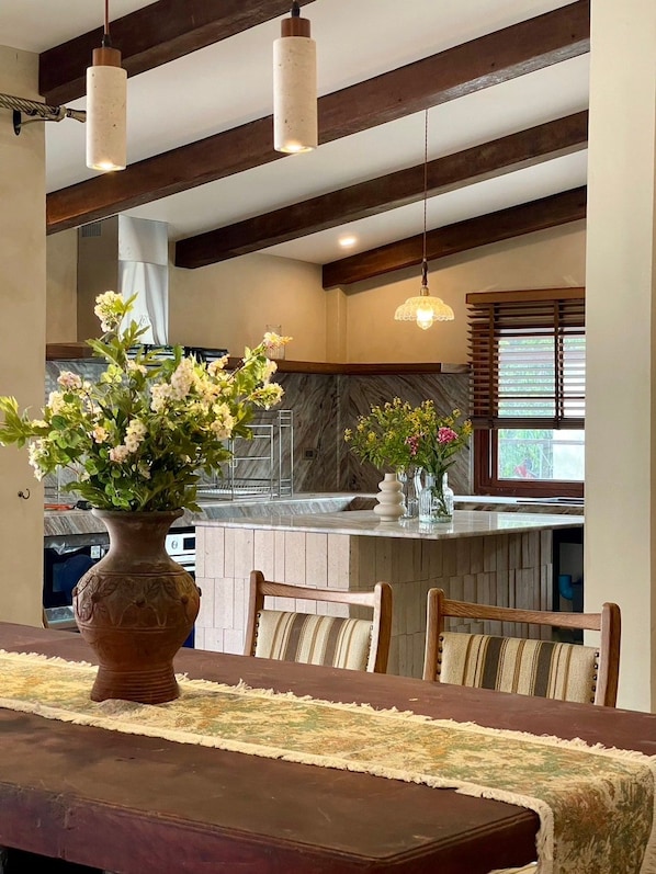 Uma sala de jantar com mesa de madeira, cadeiras, um vaso com flores e uma cozinha ao fundo.