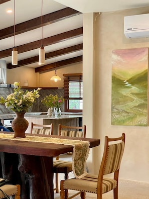 Uma área de refeições com mesa e cadeiras de madeira, um vaso com flores, luminárias pendentes e um quadro na parede.