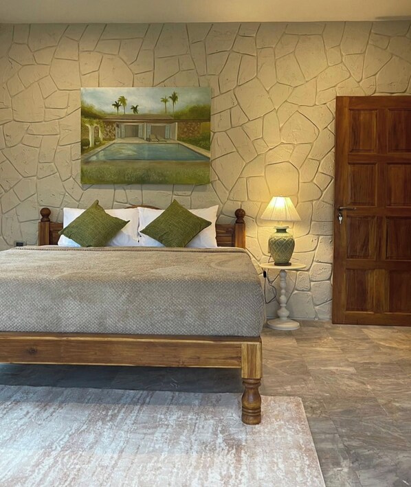 Een slaapkamer met een stenen muur, een bed met houten frame, een nachtkastje met een lamp en een deur.
