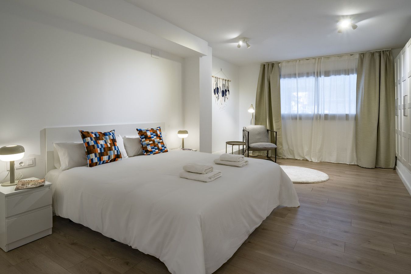 A hotel room with a large bed, bedside tables, a chair, a lamp, and a window with curtains.