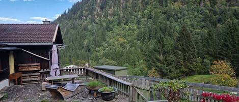 A rustic cabin with a stone foundation, wooden deck, and a fenced garden area with potted plants and flowers.