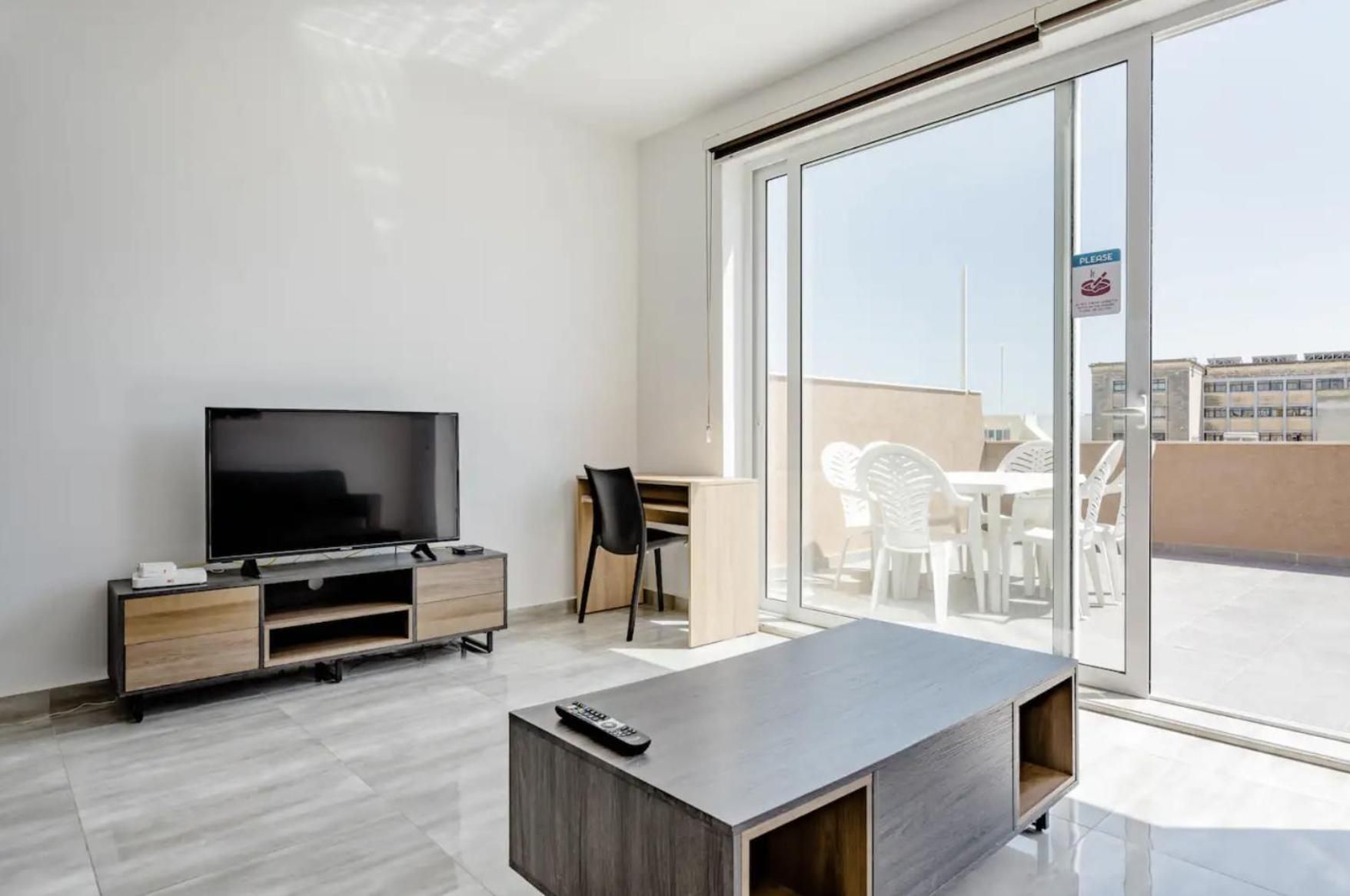 Een moderne woonkamer met een flatscreen televisie, een houten dressoir, een bureau, een stoel en een balkon met wit tuinmeubilair.