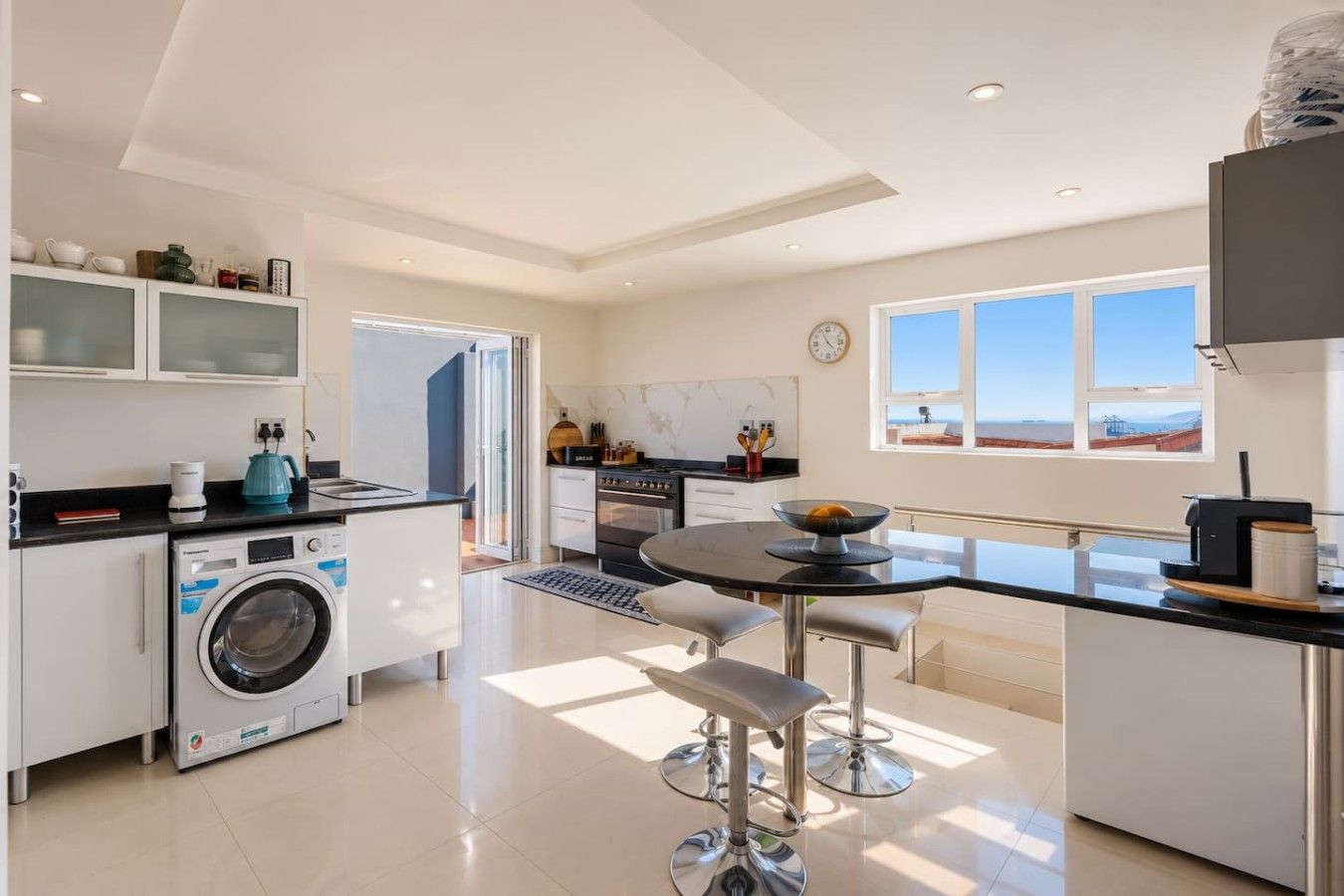 A modern kitchen with a washing machine, a dining area with a table and chairs, and a view of the sea through large windows.